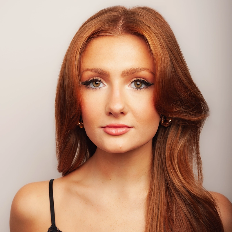 A young woman with long straight auburn hair poses in front of a neutral background. She has fair skin, hazel eyes, winged eyeliner, and natural makeup. Megan Ludwig wears gold hoop earrings and a black spaghetti-strap top, looking directly at the camera.
