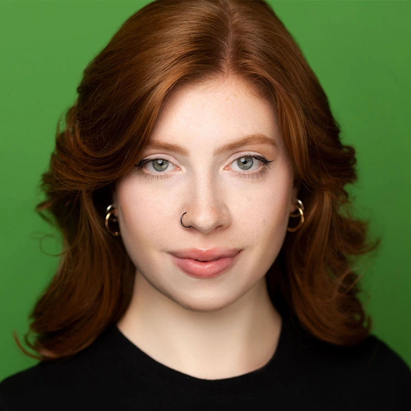 A young woman with fair skin, long wavy red hair, and light eyes looks at the camera. Jayline Ross wears hoop earrings, a nose ring, and a black top against a solid green background.