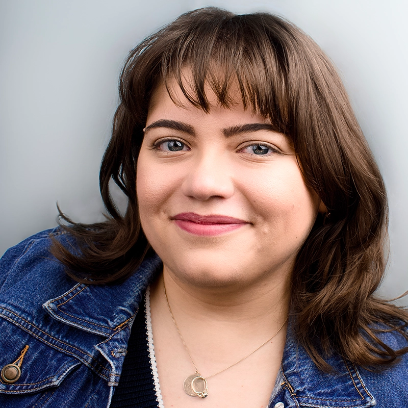 A woman with light skin, brown hair, and bangs smiles softly. She wears a blue denim jacket, a black top, and a gold necklace with a round pendant. The background is a plain light gray, complementing Michelle Zink-Muñoz’s relaxed and approachable style.