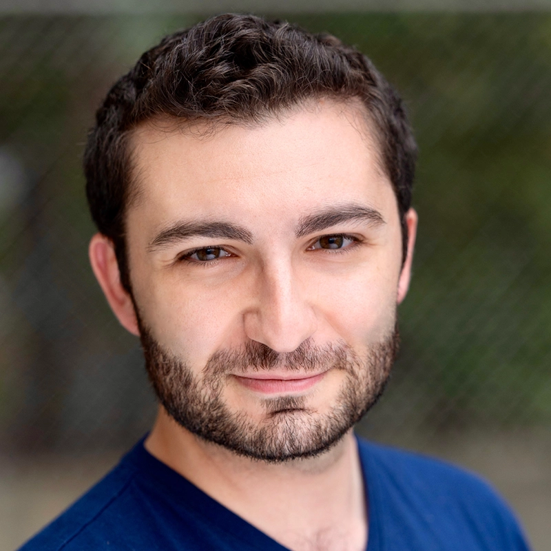 Casey Keeler, a man with short, dark hair and a full beard, smiles slightly at the camera. He wears a navy blue shirt and stands outdoors in front of a blurred green background with a wire fence. The lighting is soft and even.