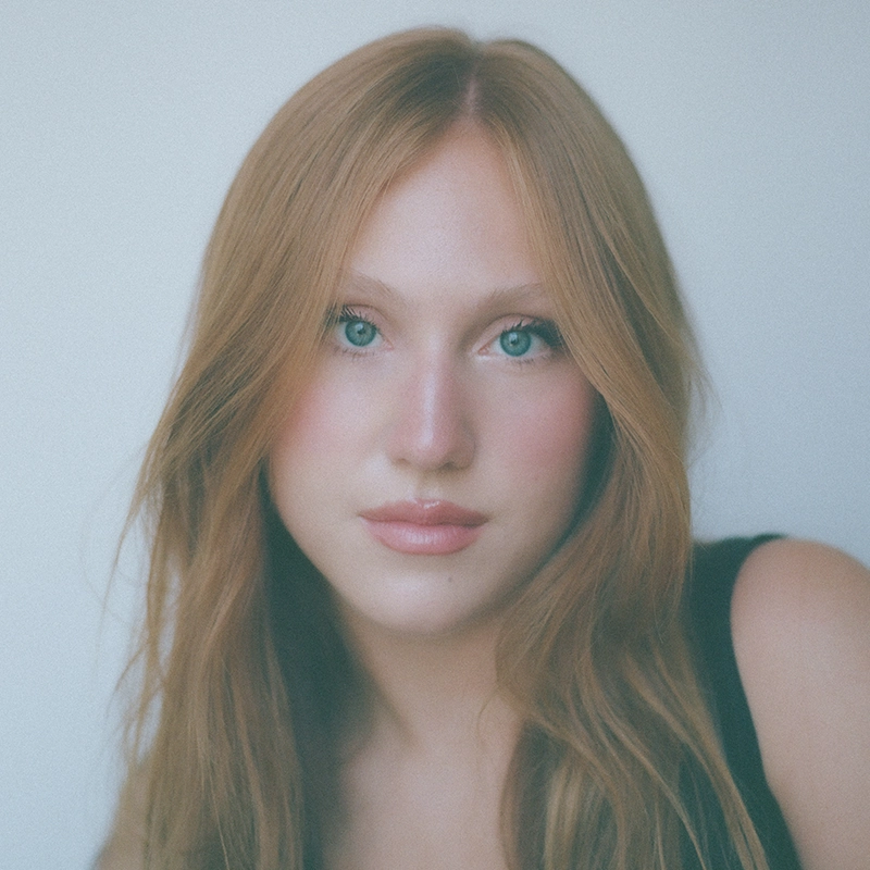 A young woman, resembling Millie Yocum, with long reddish-blonde hair and blue eyes looks at the camera. She has fair skin, natural makeup, and wears a sleeveless black top. The background is plain and softly blurred, highlighting her face with a soft and even lighting that creates a delicate surfac of luminosity on her features.