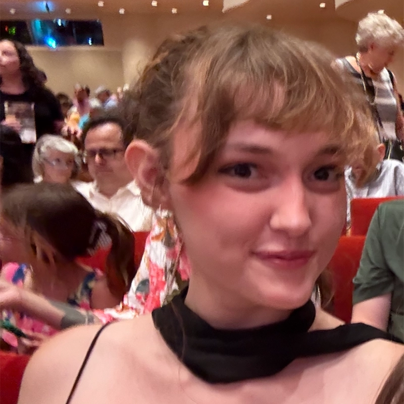 A young woman with light brown hair and bangs smiles at the camera in a theater. She wears a black sleeveless top with a scarf, capturing a moment as lively as one of Phillip Wong's stage productions. Red seats and several people, including children and adults, are visible in the background, some looking in different directions.