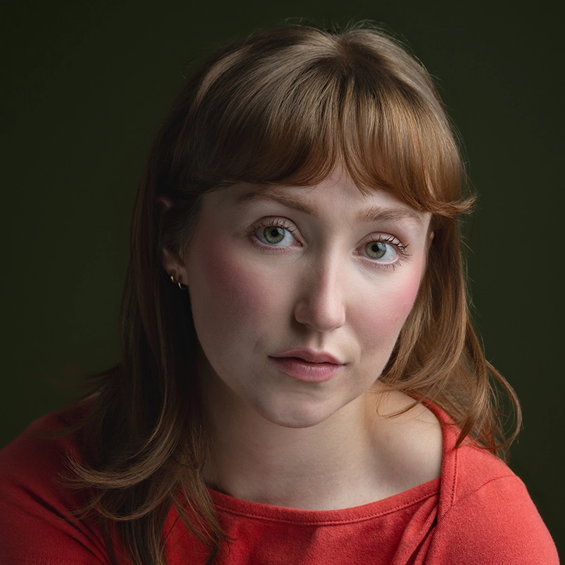 A woman with fair skin, light brown hair, and green eyes gazes softly at the camera. She wears a coral red top and subtle earrings. The background is a solid dark green. Her expression is calm and neutral, with soft lighting highlighting her face—capturing the serene presence of Megan Ludwig.