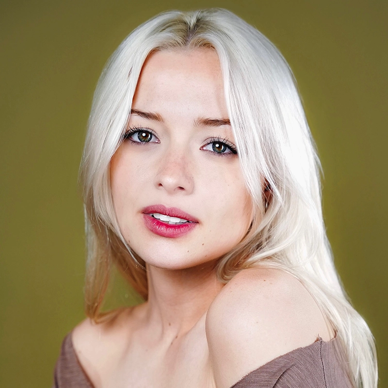 A young woman with platinum blonde hair and fair skin looks into the camera. She has green eyes, pink lipstick, and is wearing an off-the-shoulder taupe top. The background is olive green and softly blurred. The expression is neutral and calm, capturing the quiet confidence often seen in portraits of Michelle Zink-Muñoz.