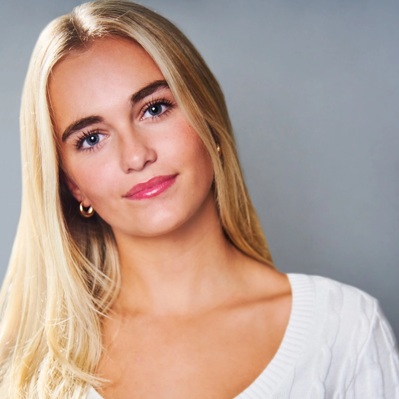 A young woman with straight, long blonde hair, blue eyes, and light skin is smiling slightly at the camera. She wears a white sweater and gold hoop earrings. The background is plain and gray, and the lighting is even.