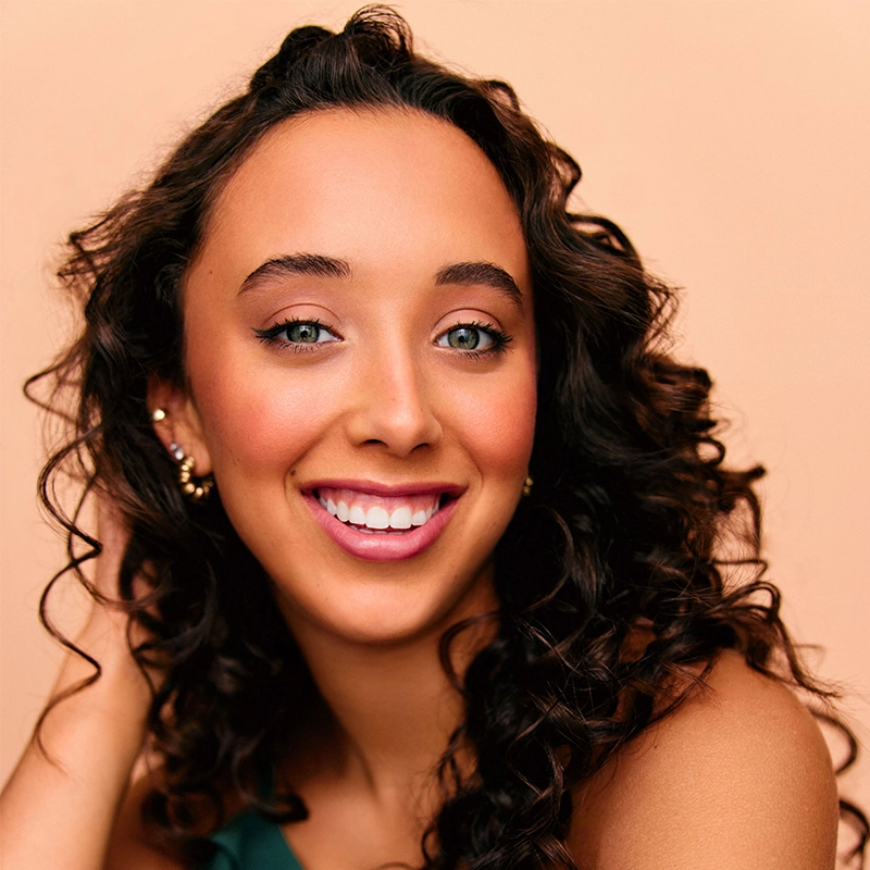 Gabrielle Farley, a young woman with long, dark curly hair, smiles brightly at the camera. She wears gold earrings and subtle makeup, including pink lipstick. Her hand touches her hair as she poses against a smooth, peach-colored background.