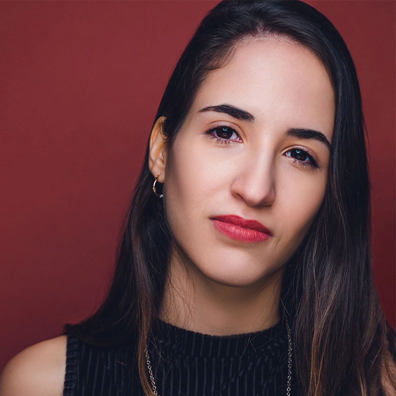 A young woman with long dark hair and light skin looks into the camera. She wears a sleeveless black top, small gold hoop earring, and a subtle necklace. Her expression is neutral. The background is a solid reddish-brown color.