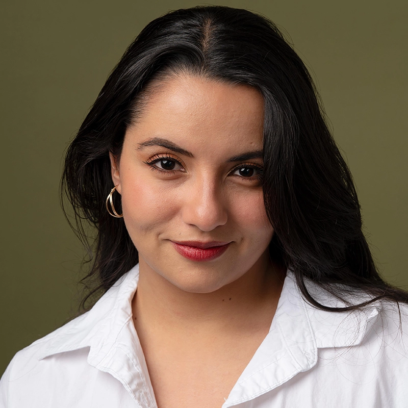 A woman with long dark hair, gold hoop earrings, and red lipstick smiles softly. She wears a white collared shirt and faces the camera against an olive green background. The lighting is even, highlighting her features and creating a professional, warm portrait.