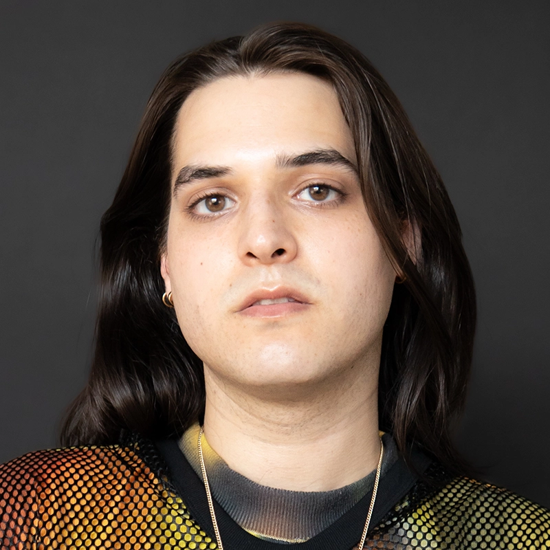 Nic Sylvester, with long dark brown hair, wearing a multicolored shirt and gold necklace, faces forward with a neutral expression. They have light skin and are set against a plain dark background. Their eyes are light brown and they wear a small hoop earring.