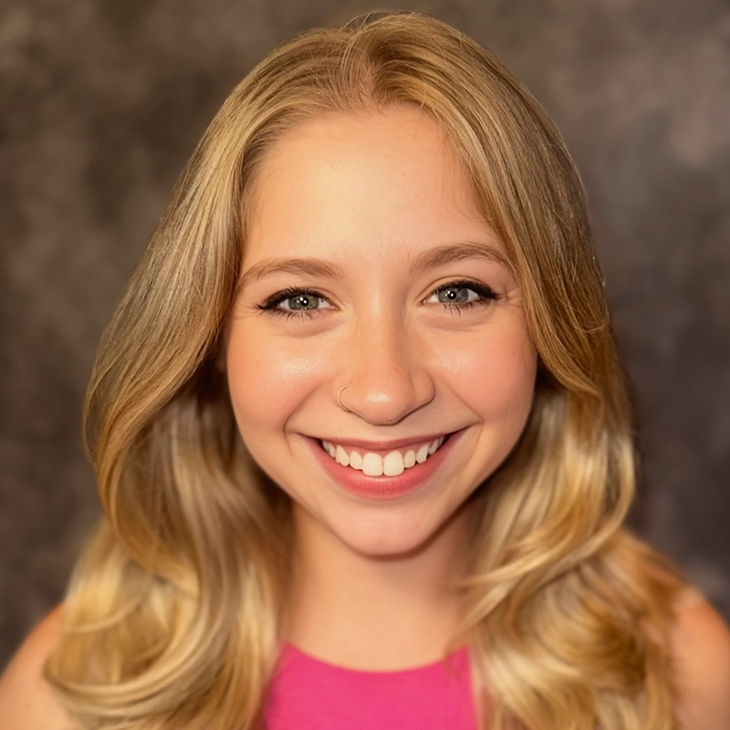 A young woman with long blonde hair, wearing a pink sleeveless top, smiles at the camera. She has a small nose ring and subtle makeup. The background is blurred and dark, highlighting Casey Keeler’s bright, friendly expression.