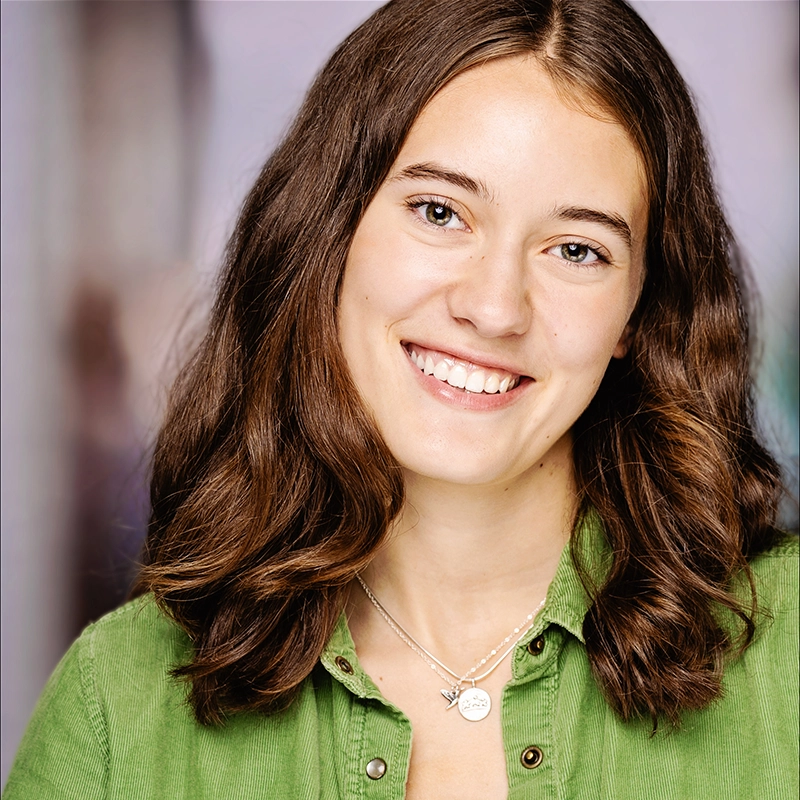 Jenny Walker, a young woman with wavy brown hair, smiles at the camera. She wears a green shirt and two silver necklaces. The background is softly blurred with neutral tones, drawing focus to her friendly expression.