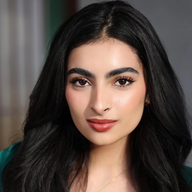 A woman with long black hair and bold eyebrows looks directly at the camera. She has winged eyeliner, rosy cheeks, and coral lipstick. The background is softly blurred with neutral tones, highlighting her clear skin and confident expression.