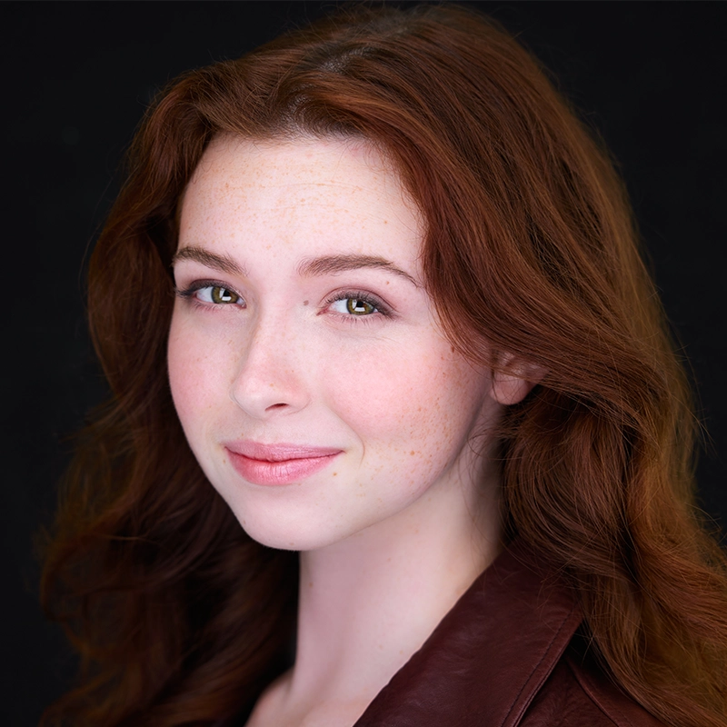 A young woman with fair skin, long wavy auburn hair, and green eyes smiles softly. She wears a brown leather jacket and light makeup. The background is plain black, making her face the focal point of the image in this striking Gaël Mouton-Sady portrait.