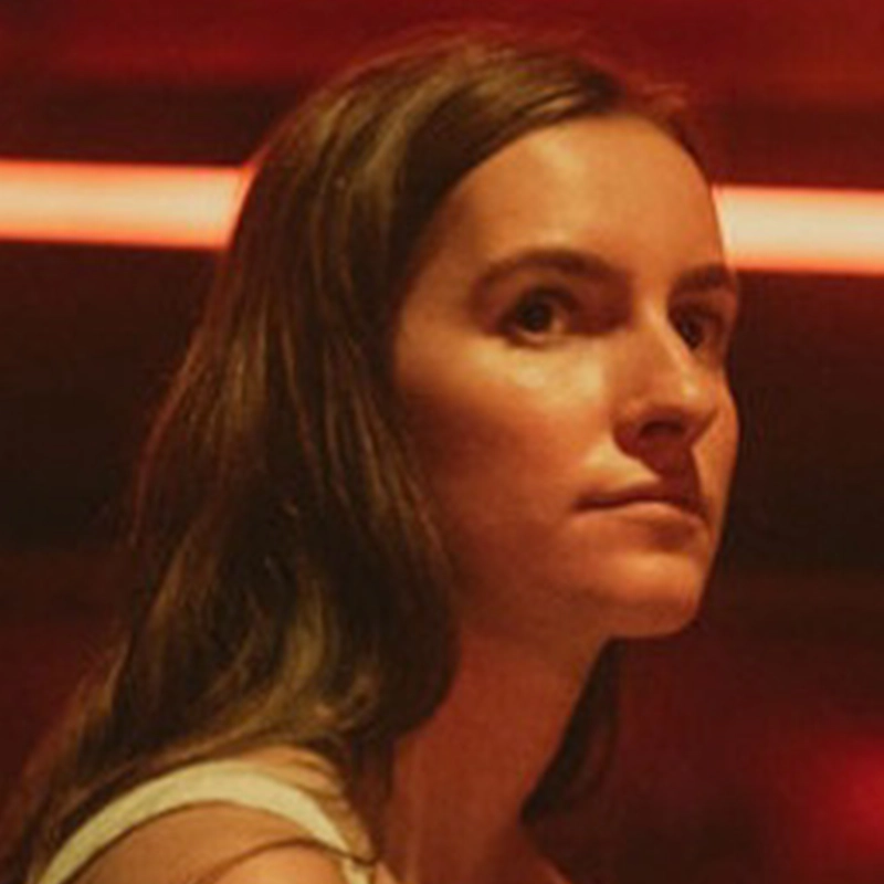 A young woman with long brown hair, identified as Olivia Kelly, looks to the side under warm, reddish lighting. She has a serious expression and is shown from the shoulders up, with a blurred red horizontal light in the background.