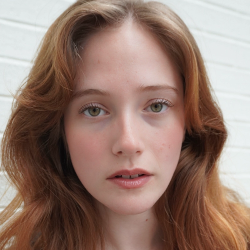 A young woman with light skin and long, wavy auburn hair stares directly at the camera. She has green eyes, natural makeup, and a neutral expression. A white brick wall serves as the background, creating a minimalist setting reminiscent of Asher Alcantara’s signature portrait style.