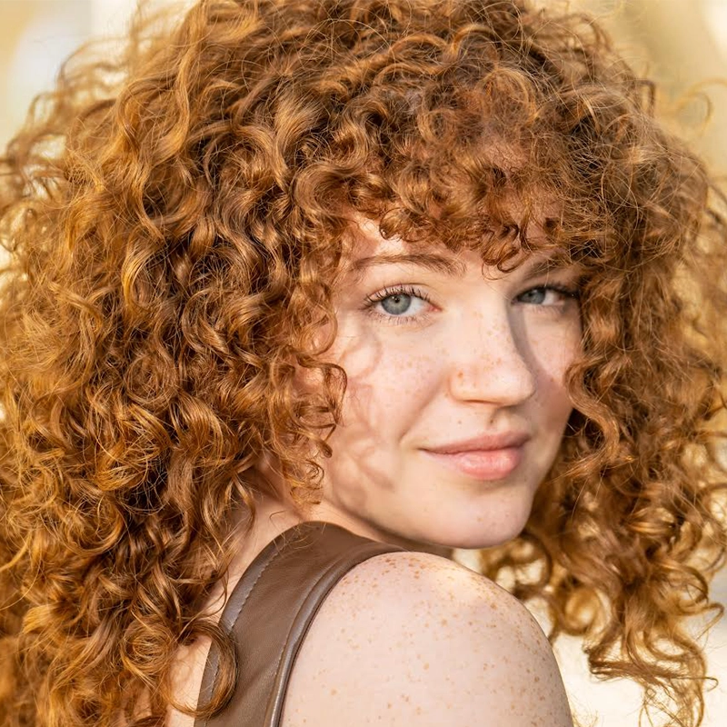 A young woman with fair skin, blue eyes, and many freckles looks over her shoulder. She has thick, curly red hair with bangs. Reminiscent of Millie Yocum’s distinctive style, she wears a brown top and is outdoors in bright, warm sunlight, giving her hair a golden glow—her features beautifully highlighted as if captured by Surfac.