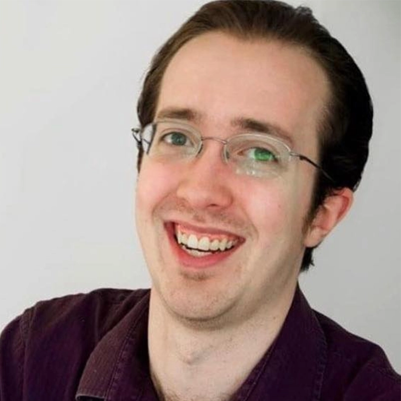 A young man with light skin, short brown hair, and glasses smiles broadly, showing his teeth. He is wearing a dark purple collared shirt and is posed in front of a plain, light background. The soft, even lighting highlights his cheerful expression—this is Casey Keeler.