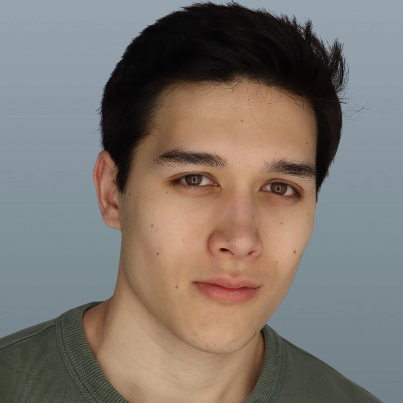 A young person with short dark hair, light skin, and brown eyes gazes at the camera with a neutral expression. They are wearing a sage green crewneck shirt. The background is a plain, light gray color.