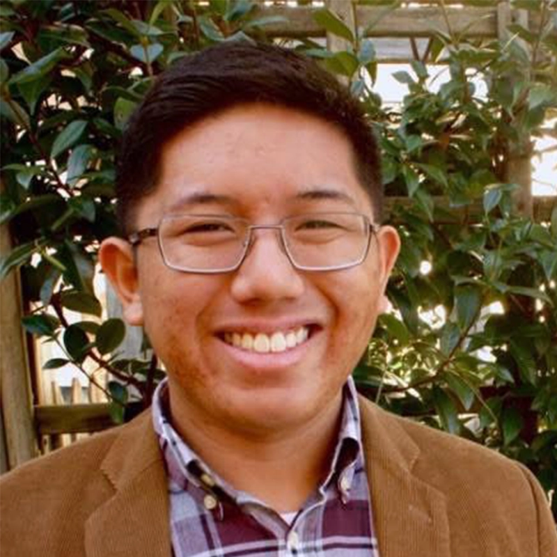 Phillip Wong, a young person with short dark hair and glasses, smiles outdoors, wearing a brown jacket over a plaid shirt. Green leaves and a wooden trellis fill the background under natural light.