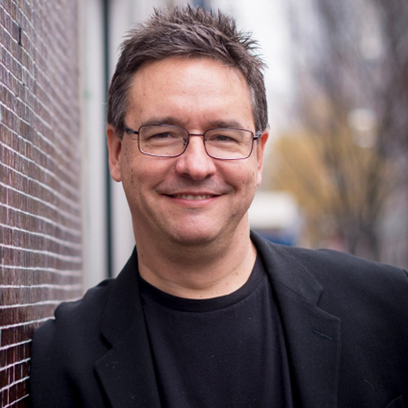 A man with short, spiked dark hair and glasses smiles at the camera. He is wearing a black blazer over a black shirt and is standing outside, leaning casually against a brick wall. The background appears blurred with hints of trees and buildings.