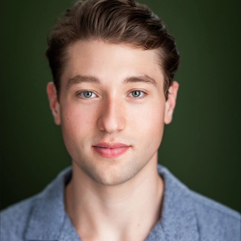 Young man with fair skin, short brown hair, and blue eyes looks straight at the camera with a neutral expression. Jack Plogger wears a light blue collared shirt. The background is a solid, dark green color. Image is well-lit, highlighting his facial features.
