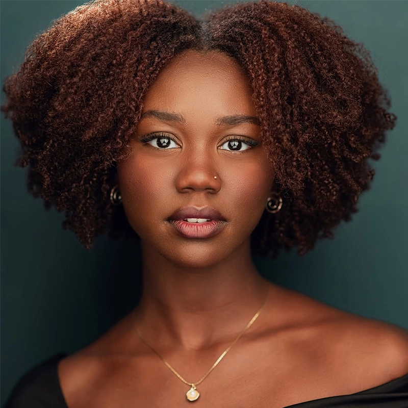 A young woman with medium brown skin and natural, voluminous curly hair poses against a dark green background. Wearing a black off-shoulder top and gold necklace, she embodies Qawiyya—her calm, confident expression radiates strength and self-assurance.