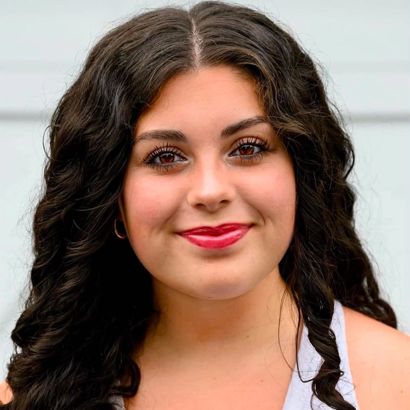 A young woman with long, dark curly hair smiles gently. She has brown eyes, bold eyebrows, glowing skin, and wears bright red lipstick. Laney Rakofsky is dressed in a white sleeveless top, and the background is a plain light gray wall.