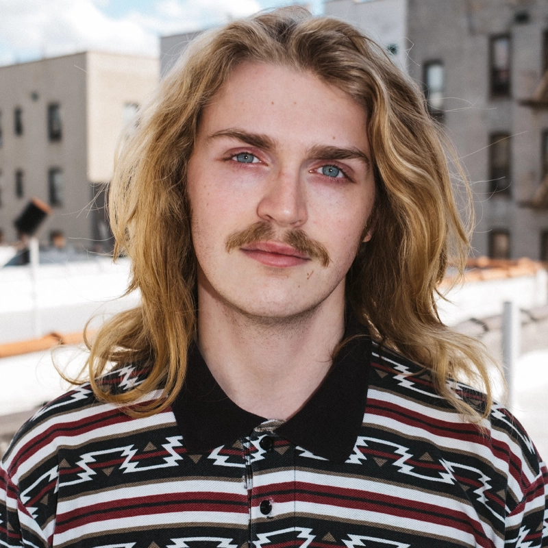A young adult with long blonde hair and a mustache stands outside. He wears a patterned shirt with black, white, and red zigzags. The background shows blurred buildings and a bright, partly cloudy sky, capturing Mitchell Reed in a candid urban moment.