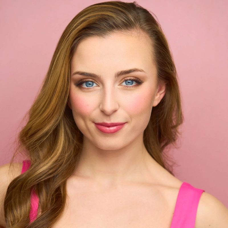 Rachel Revellese, a young woman with long, wavy light brown hair and blue eyes, smiles slightly. She wears a sleeveless pink top and natural makeup with pink blush and lipstick. The background is a solid pink color.
