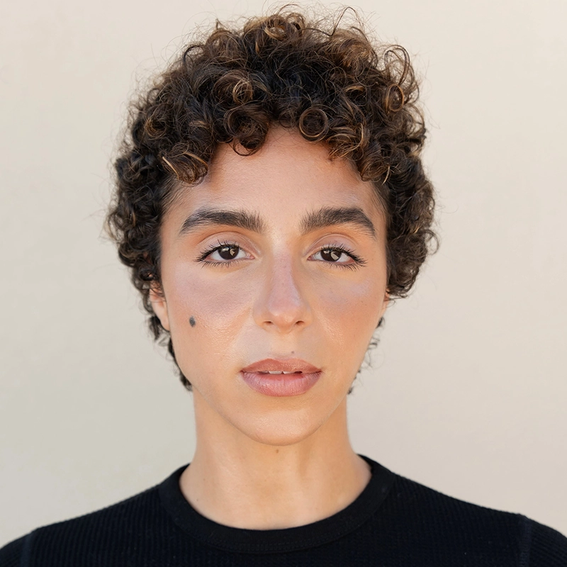 A person with short, dark, curly hair and thick eyebrows looks directly at the camera. They have light skin, a prominent mole on their left cheek, and wear a black top. The background is plain and neutral, drawing focus to their face.