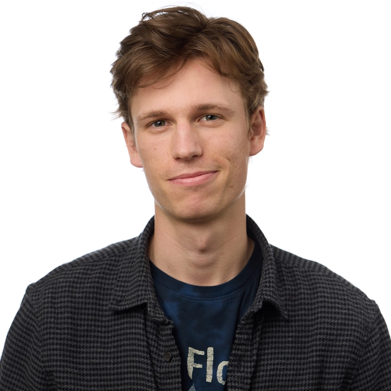 A young man, Izaiah Rhinehart, with light brown hair and fair skin smiles softly. He wears a dark checkered shirt over a navy blue graphic tee. The background is plain white, and the lighting is bright and even. His hair is tousled, and he looks relaxed and approachable.