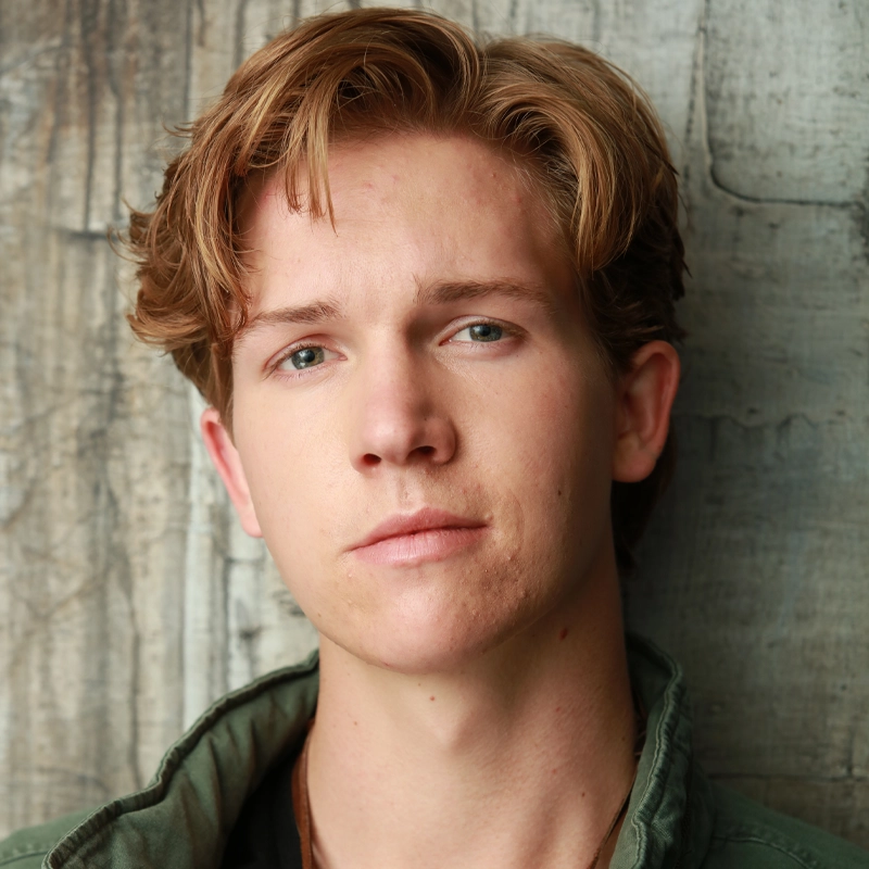 A young person with light skin, short, wavy, reddish-brown hair, and blue eyes looks into the camera with a neutral expression. Wyatt Rhinehart wears a green jacket and stands against a weathered wooden wall.