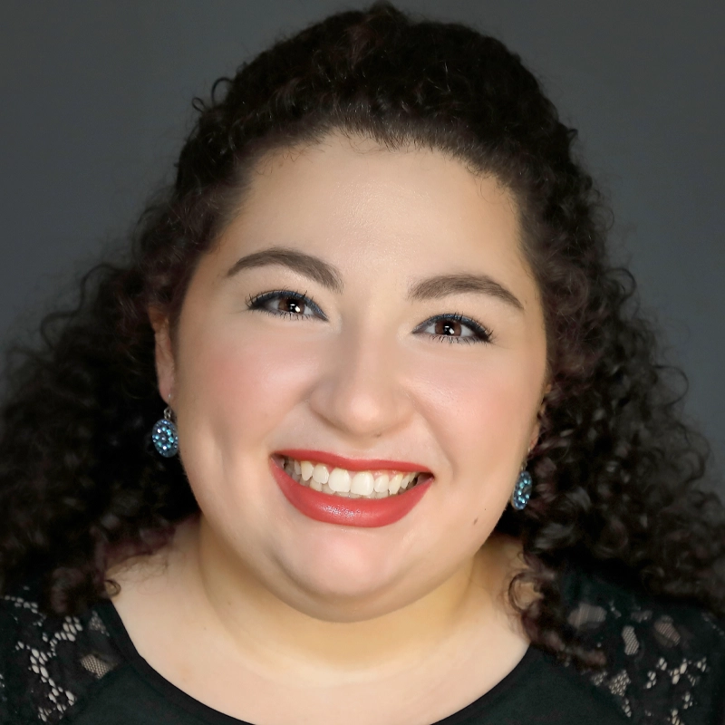 Ariel Richter, a woman with curly dark hair, smiles widely, showing her teeth. She wears red lipstick, blue earrings, and a black top with lace details on the shoulders. The background is plain and dark, highlighting her face.