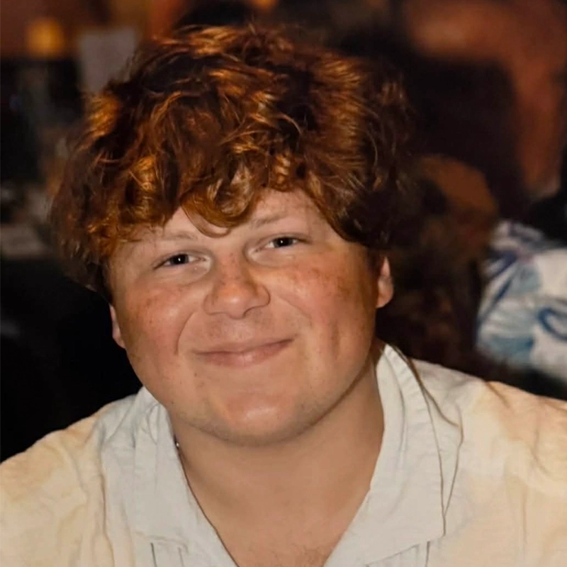 A young person with wavy red-brown hair and fair skin smiles softly. They wear a light-colored shirt. The background is blurred with dark and warm tones, suggesting an indoor, possibly social setting—perhaps somewhere Phillip Wong might feel at home.