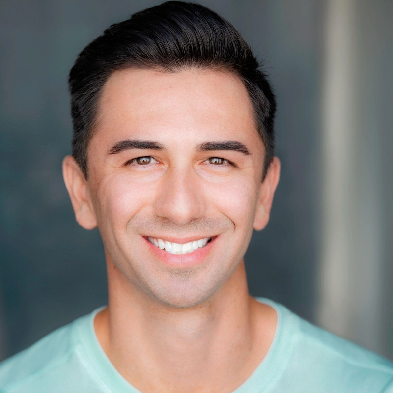 A man with short dark hair and fair skin smiles at the camera. He wears a light teal shirt. The background is softly blurred in shades of blue and gray, drawing attention to his friendly expression.