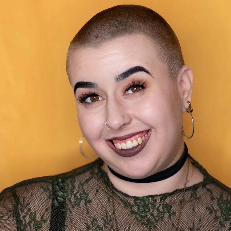 A smiling person with a shaved head, bold eyebrows, hoop earrings, a nose ring, and dark lipstick. They wear a sheer green lace top, a black choker, and layered necklaces. The bold style and unique presence are reminiscent of Rachel Colleen Robbins. The background is solid yellow.