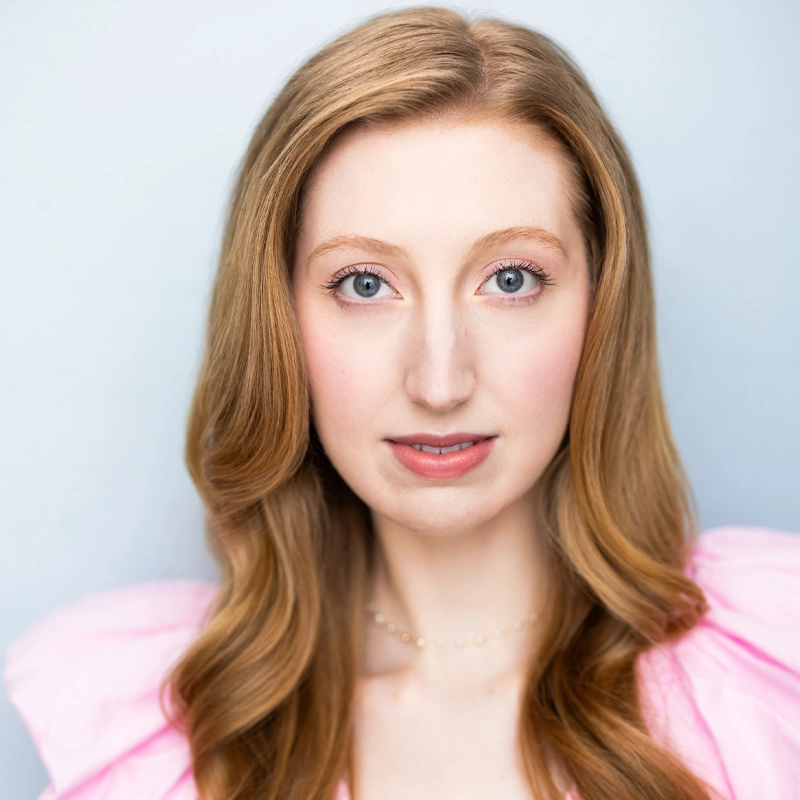 A woman with long, wavy light brown hair and blue eyes looks at the camera. She wears a light pink, ruffled top and a delicate pearl necklace. The background is a soft, light blue. The lighting is bright and even, highlighting her features.