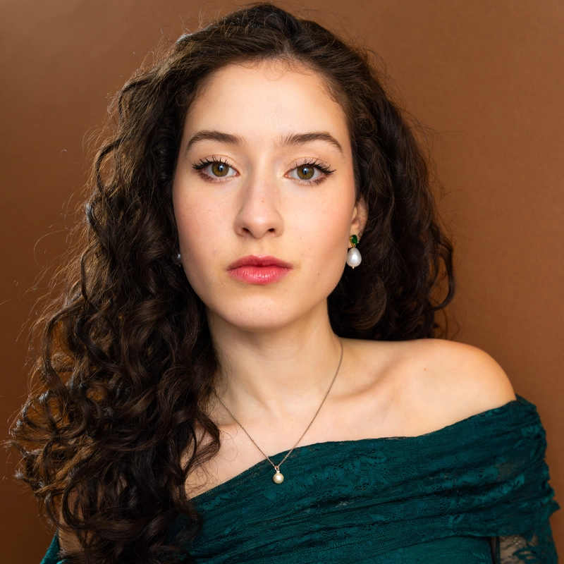 A young woman, Anjali Rooney, with long, dark curly hair, light skin, and hazel eyes looks at the camera. She wears a green off-shoulder top, pearl earrings, and a necklace with a small pendant. The background is plain brown. Her expression is neutral.