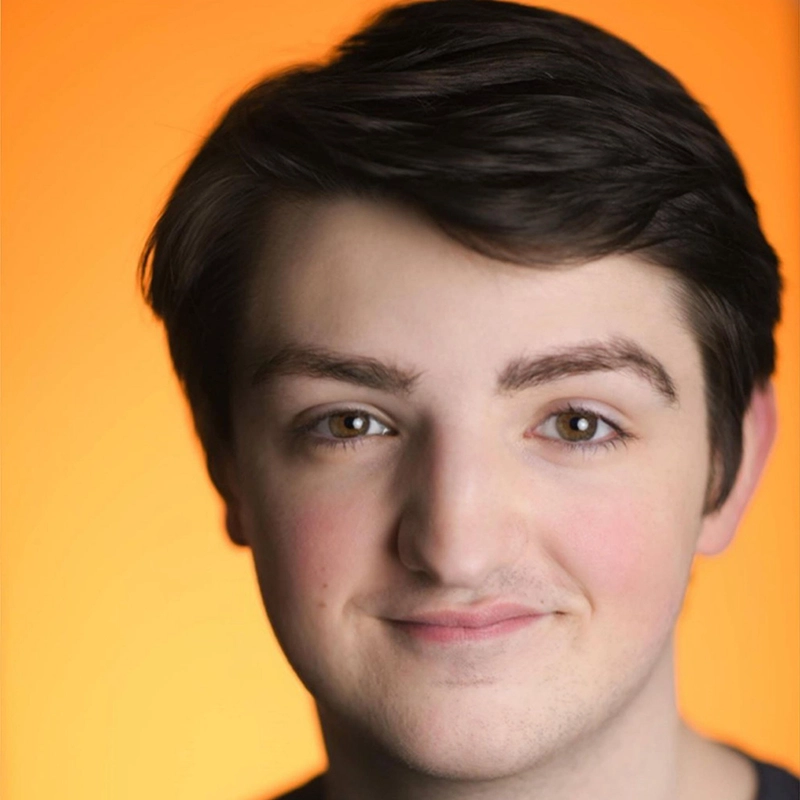 A young person with short brown hair and fair skin smiles gently. Their expression is friendly, and they are facing the camera. The background is a smooth, warm orange gradient. Their eyes are brown and eyebrows are dark, capturing a candid, approachable moment reminiscent of Gabrielle Farley’s warm presence.
