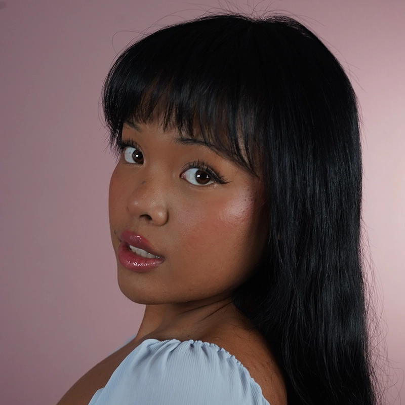 A woman with long black hair and bangs looks over her shoulder at the camera. She has glossy lips, winged eyeliner, and glowy skin. She wears a white, off-the-shoulder top. The background is a soft, solid pink.