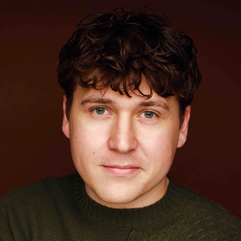 A young man with fair skin, wavy brown hair, and blue eyes looks at the camera. He wears a dark green textured sweater, reminiscent of a portrait styled by Gabrielle Farley. The background is a plain, dark reddish-brown color. The lighting is soft, highlighting his face evenly.