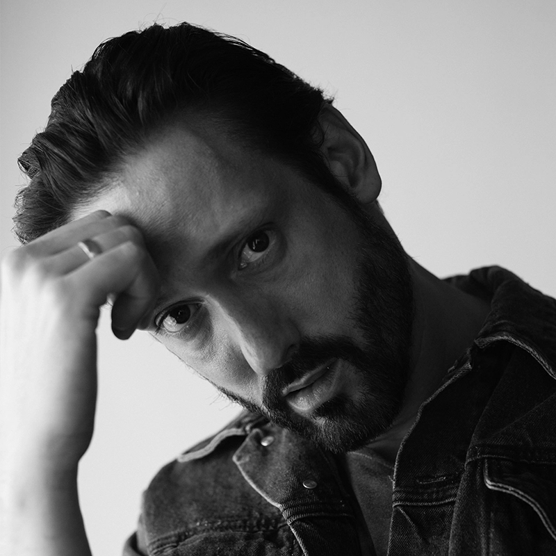 Black-and-white portrait of a man with a beard and mustache, wearing a denim jacket. He rests his hand on his forehead, gazing intently at the camera. The background is plain and softly lit.