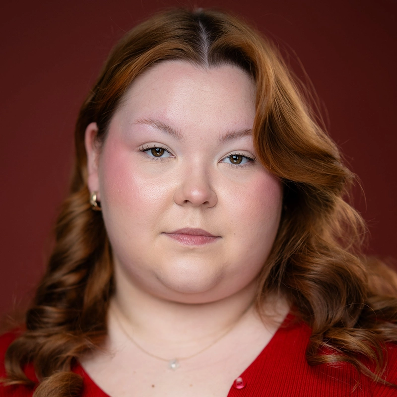 A woman with fair skin and wavy auburn hair poses against a reddish-brown background. She wears a red top, small hoop earrings, and a subtle necklace. Her makeup is natural, with pink blush and light lipstick, and she looks calmly at the camera—perfectly capturing Megan Ludwig’s understated elegance.