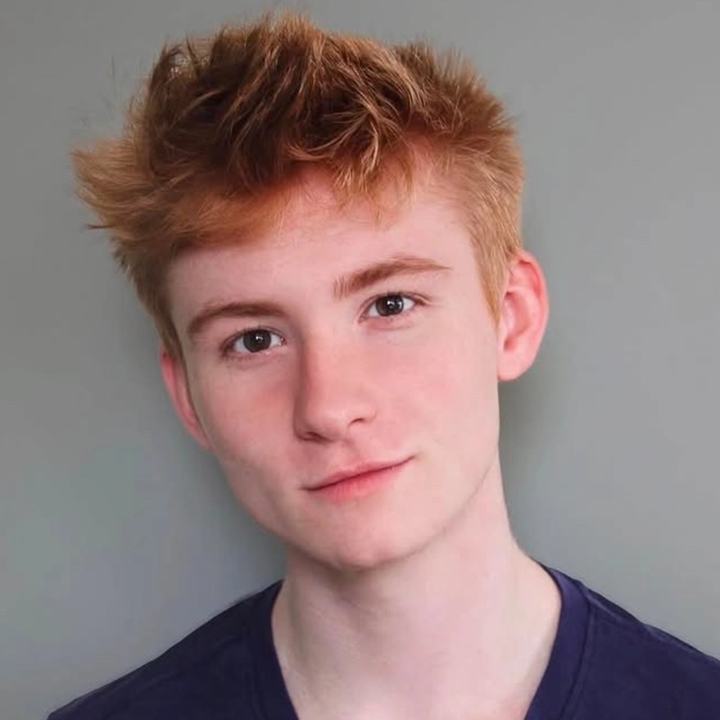 A young person with messy red hair and fair skin looks at the camera with a slight smile. They are wearing a dark blue shirt and standing against a plain gray background. The lighting is soft and even across their face.