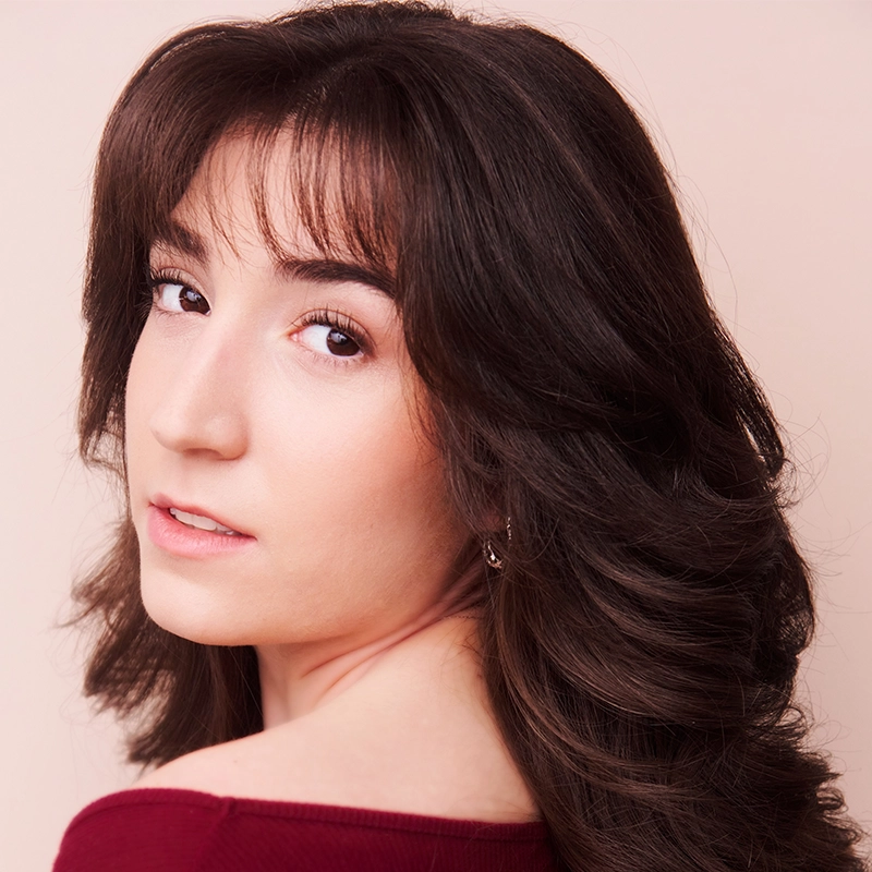A young woman with wavy brown hair and side-swept bangs looks over her shoulder at the camera. Saylor Tucker wears a maroon top, has light makeup, and a soft, neutral expression against a pale, solid background.