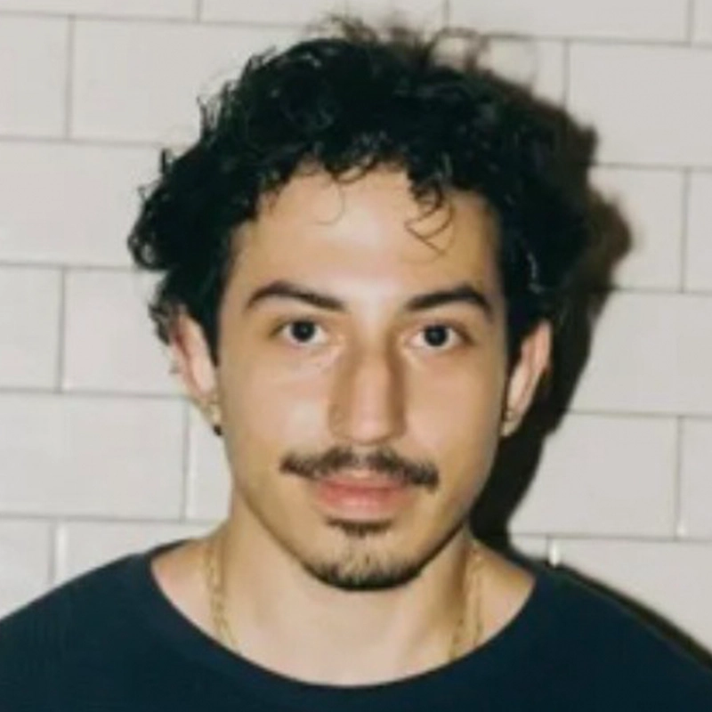 A person with short curly dark hair, a mustache, and light skin faces the camera. They are wearing a dark shirt, gold necklace, and small hoop earrings. The background consists of white subway tiles. This individual is Samuel Rossi.