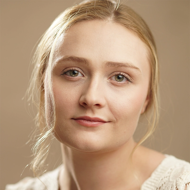 A young woman with fair skin, blond hair pulled back, and green eyes looks into the camera with a relaxed, slight smile. She wears a cream knit sweater. The background is soft beige, emphasizing her calm expression and natural makeup—an effortlessly elegant portrait of Alexandra Zografos.