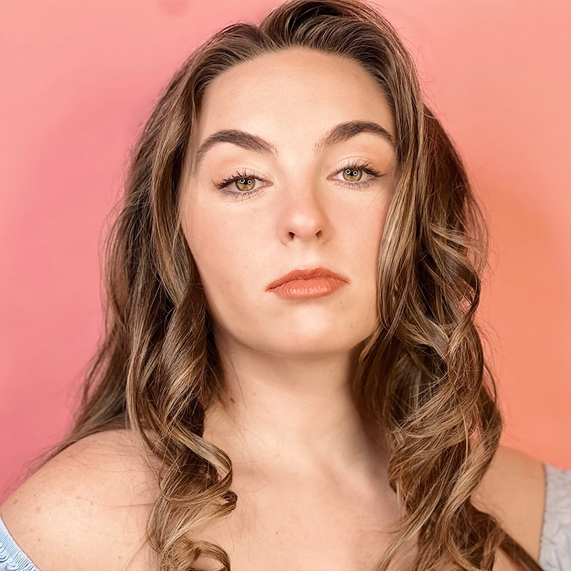 A woman with wavy brown hair poses against a pink and orange gradient background in a portrait by Phillip Wong. She has light skin, arched eyebrows, and wears natural makeup with glossy lips. Her expression is neutral and she wears an off-the-shoulder light blue top.
