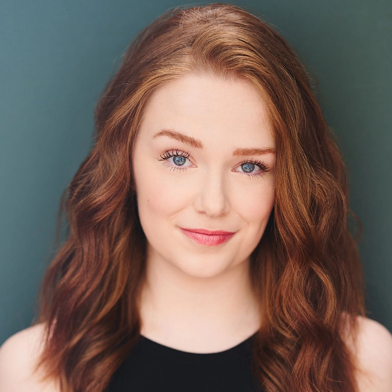 A young woman, resembling Millie Yocum Surfac, with wavy auburn hair, blue eyes, and fair skin looks at the camera against a teal background. She wears a sleeveless black top and has a slight smile, with natural makeup and well-groomed eyebrows.