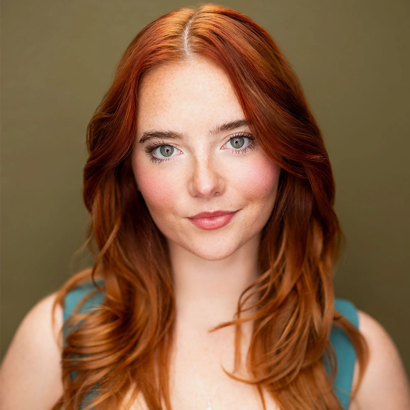 A woman with long, wavy red hair and fair skin is centered against a plain olive background. She has blue eyes, subtle makeup, and wears a teal sleeveless top. Looking directly at the camera with a soft, confident expression, Saylor Tucker exudes quiet poise and self-assurance.
